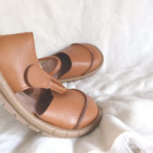 May include: A pair of tan leather shoes with a pebbled texture, featuring a rounded toe, a T-strap design with a circular closure, and a thick, ridged rubber sole. The shoes have a visible seam detail across the front and a smooth interior lining.