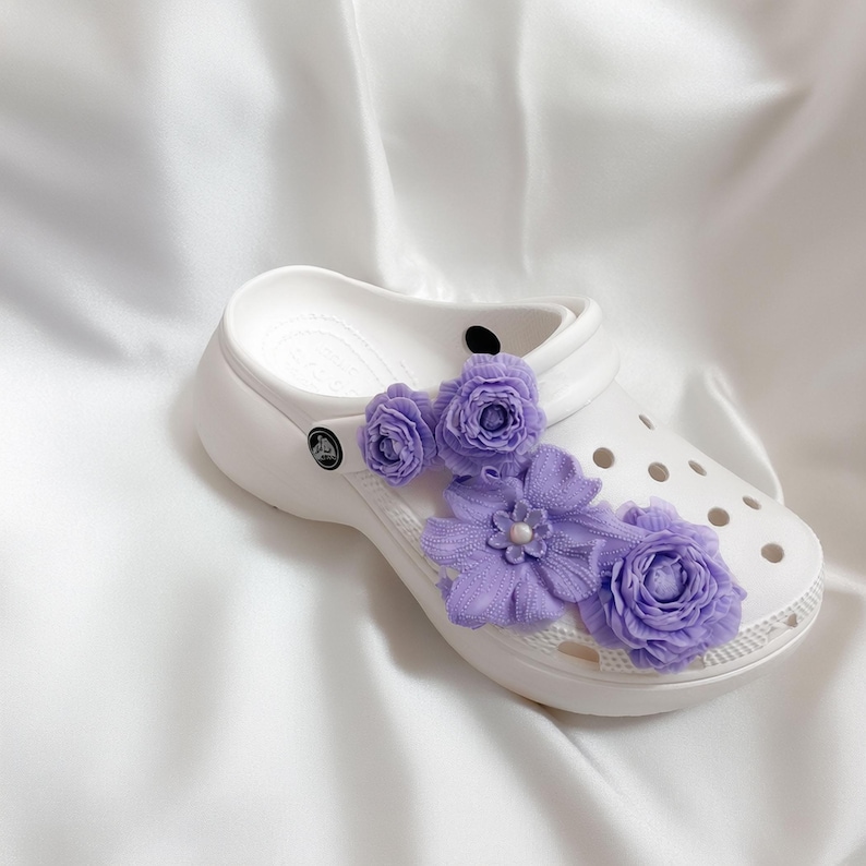 Pu&ograve; includere: Zoccoli bianchi adornati con delicati fiori di tessuto lavanda. Gli abbellimenti floreali includono diversi piccoli boccioli a forma di rosa e un gruppo pi&ugrave; grande con un centro di perle, aggiungendo un tocco di eleganza alle calzature casual.