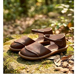 May include: A pair of brown leather sandals featuring a rounded closed-toe design, a textured pebbled surface, and a single ankle strap with a side closure. The sandals have a flat, flexible sole and a smooth interior lining.