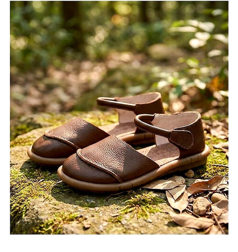 May include: A pair of brown leather sandals featuring a rounded closed-toe design, a textured pebbled surface, and a single ankle strap with a side closure. The sandals have a flat, flexible sole and a smooth interior lining.