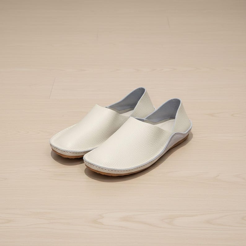May include: A pair of off-white leather slip-on shoes. The shoes have a rounded toe, a low profile, and a light brown sole. The upper is a textured leather, and the interior is a light blue color. The shoes are designed for comfort and casual wear.