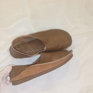 May include: A pair of brown leather slippers. The slippers have a rounded toe and a low profile. The interior is a lighter brown color. The sole is a darker brown. The slippers are made of soft leather.