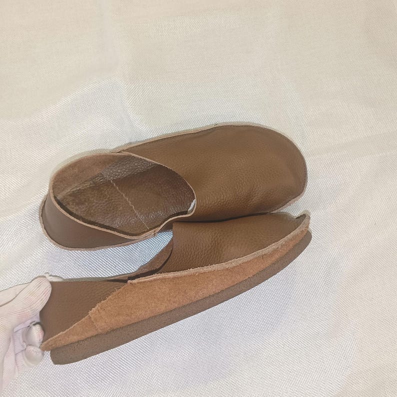 May include: A pair of brown leather slippers. The slippers have a rounded toe and a low profile. The interior is a lighter brown color. The sole is a darker brown. The slippers are made of soft leather.