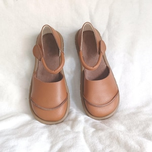 May include: A pair of tan leather shoes with a rounded toe, a textured pebble-grain finish, and a single ankle strap with a side closure. The shoes feature a low-profile design with a smooth inner lining and a flexible, flat rubber sole.