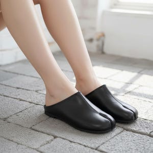 Op de afbeelding: Zwarte leren instappers met split-teen. De schoenen hebben een minimalistisch design met een ronde neus en een lage hak. Het split-teenontwerp is een uniek kenmerk en de schoenen lijken van glad leer te zijn.