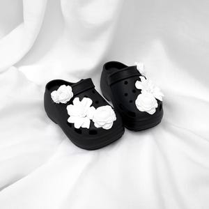 Pu&ograve; includere: Un paio di zoccoli neri con plateau adornati da decorazioni floreali bianche. Gli zoccoli hanno un design classico con cinturino e fori di ventilazione. I fiori bianchi sono raggruppati sulla parte superiore e sui lati delle scarpe.