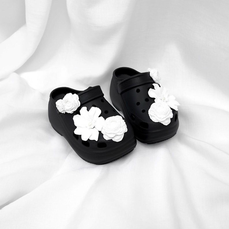 Pu&ograve; includere: Un paio di zoccoli neri con plateau adornati da decorazioni floreali bianche. Gli zoccoli hanno un design classico con cinturino e fori di ventilazione. I fiori bianchi sono raggruppati sulla parte superiore e sui lati delle scarpe.