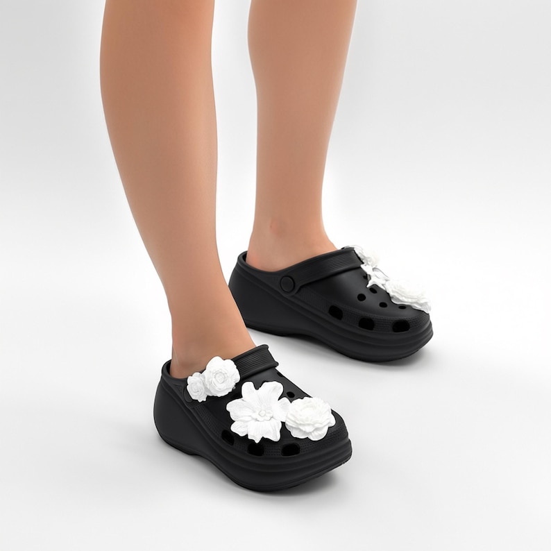 Pu&ograve; includere: Zoccoli neri con decorazioni floreali bianche. Questi zoccoli hanno una suola platform e fori di ventilazione. I fiori bianchi sono raggruppati sulla parte superiore, aggiungendo un tocco decorativo alle calzature.