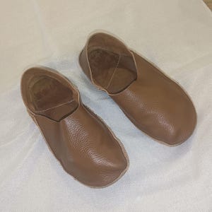 May include: A pair of brown leather slippers. The slippers have a rounded toe and a low-cut design, exposing the top of the foot. The leather has a textured appearance. The slippers are likely for indoor use.