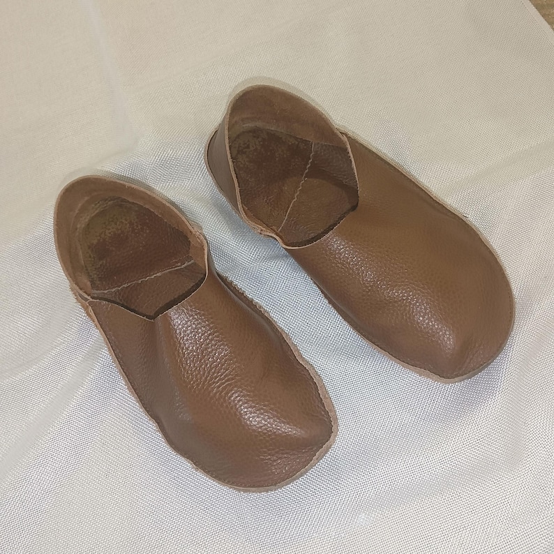 May include: A pair of brown leather slippers. The slippers have a rounded toe and a low-cut design, exposing the top of the foot. The leather has a textured appearance. The slippers are likely for indoor use.