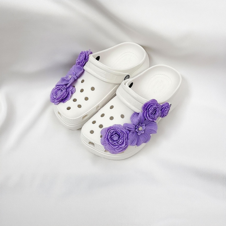 Pu&ograve; includere: Un paio di zoccoli bianchi adornati con decorazioni floreali viola. Gli zoccoli presentano un design classico con molteplici fori di ventilazione e un cinturino. I fiori viola sono raggruppati sui lati, aggiungendo un tocco decorativo alle calzature.