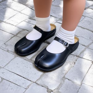 May include: Black leather split-toe clogs with a strap and buckle closure. The clogs are worn with white socks. The shoes have a unique design, with a separate compartment for the big toe. The clogs are on a stone surface.