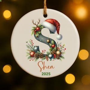 May include: A round Christmas ornament with a gold string. The ornament features the name "Shea" and the year "2025". The letter "S" is decorated with a Santa hat, antlers, and festive greenery.
