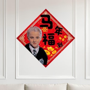 以下が含まれることがあります： 明るい髪と白黒のスーツを着た人物の肖像画が描かれた、赤いひし形の壁掛け。中国の文字と金貨も表示されています。装飾的な枕が付いた白いソファの上に設置されています。