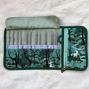 Peut inclure: Un étui à aiguilles à tricoter en tissu dans des tons verts avec un imprimé forêt, des cerfs et des arbres. L'intérieur comporte plusieurs fentes contenant des aiguilles en métal argenté, marquées de tailles métriques, et une poche zippée.