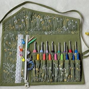 May include: An olive green fabric roll-up organiser featuring embroidered white and yellow wildflower patterns. It includes a white lace trim, a zippered pocket, and multiple slots containing various colourful metal crochet hooks.