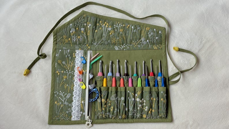 Puede incluir: Un organizador de tela enrollable en color verde oliva con patrones bordados de flores silvestres en blanco y amarillo. Incluye un borde de encaje blanco, un bolsillo con cremallera y m&uacute;ltiples ranuras con ganchillos de metal de colores.