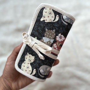 May include: A fabric roll-up organizer featuring a black background with a floral pattern and various cat silhouettes in cream, brown, and grey tones. The organizer is edged in cream-colored fabric and secured with a matching cream fabric tie.