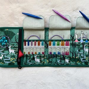 Peut inclure: Un étui de rangement en tissu vert avec un imprimé forêt et cerfs, contenant plusieurs aiguilles à tricoter et crochets colorés. L'intérieur présente des fentes étiquetées pour les tailles, une poche zippée et trois rabats supérieurs.