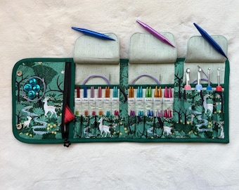 Puede incluir: Un estuche organizador de tela verde con estampado de bosque y ciervos, que contiene múltiples agujas de tejer y ganchillos coloridos. El interior tiene ranuras etiquetadas para tamaños, un bolsillo con cremallera y tres solapas superiores.