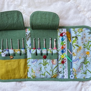 May include: A green fabric organizer case featuring two top flaps, multiple interior pockets, and a zippered compartment. The case holds several metal crochet hooks, colorful plastic stitch markers, and small tools against a floral print fabric.