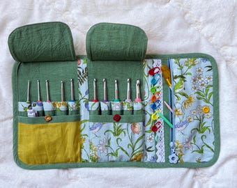 Peut inclure: Un étui de rangement en tissu vert avec deux rabats supérieurs, plusieurs poches intérieures et un compartiment zippé. L'étui contient plusieurs crochets en métal, des marqueurs de mailles en plastique colorés et de petits outils.