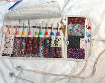 Peut inclure: Un organisateur en tissu enroulable avec un extérieur rayé crème et blanc et un intérieur en patchwork floral. Il contient plusieurs crochets aux manches colorés, une paire de ciseaux rose clair, une pochette zippée et des marqueurs de mailles.