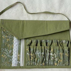 Puede incluir: Un organizador de tela enrollable en color verde oliva con cierre de solapa y cintas largas. El interior incluye un bolsillo con cremallera y encaje blanco, adem&aacute;s de una serie de ranuras verticales decoradas con motivos florales bordados.