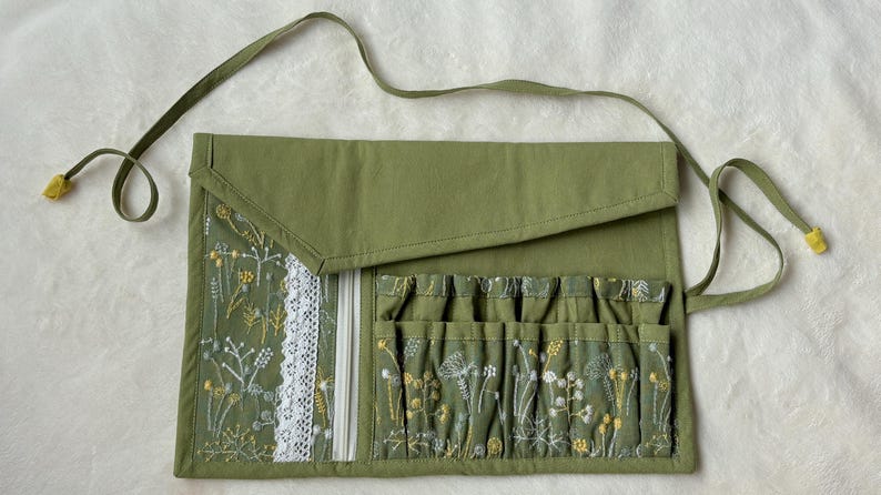 Puede incluir: Un organizador de tela enrollable en color verde oliva con cierre de solapa y cintas largas. El interior incluye un bolsillo con cremallera y encaje blanco, adem&aacute;s de una serie de ranuras verticales decoradas con motivos florales bordados.