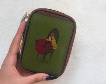 Puede incluir: Un estuche rectangular marrón con cremallera que presenta un panel frontal verde con una ilustración impresa de una figura de cabello rubio largo y vestido rojo, sentada en una silla de respaldo alto de color verde oscuro.