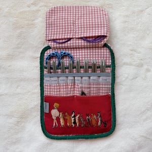 May include: A fabric knitting needle organiser case featuring a pink and white gingham pattern with a dark green trim. The case includes a red pocket with a graphic illustration of a person and several cats, holding metal needles and blue stitch markers.