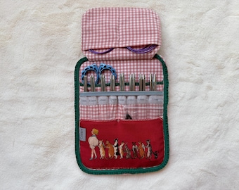 Peut inclure: Un étui de rangement en tissu pour aiguilles à tricoter avec un motif vichy rose et blanc et une bordure vert foncé. L'étui comprend une poche rouge avec une illustration graphique d'une personne et plusieurs chats, contenant des aiguilles en métal.
