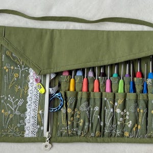 Puede incluir: Un organizador de tela verde oliva con un patr&oacute;n de bordado floral blanco y un borde de encaje blanco. Contiene m&uacute;ltiples ranuras que albergan varios ganchillos coloridos, una cinta m&eacute;trica blanca y una herramienta de pl&aacute;stico azul.
