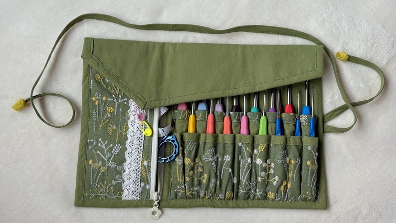 Puede incluir: Un organizador de tela verde oliva con un patr&oacute;n de bordado floral blanco y un borde de encaje blanco. Contiene m&uacute;ltiples ranuras que albergan varios ganchillos coloridos, una cinta m&eacute;trica blanca y una herramienta de pl&aacute;stico azul.