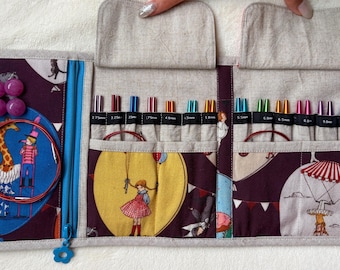 Peut inclure: Un étui de rangement en tissu pour aiguilles à tricoter avec un fond violet orné d'illustrations de personnages et d'animaux. L'intérieur comporte plusieurs compartiments contenant des aiguilles métalliques colorées avec leurs tailles en millimètres.