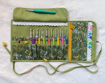 Peut inclure: Un étui de rangement en tissu vert avec un motif floral, une bordure en dentelle blanche et une fermeture éclair jaune. L'étui contient plusieurs crochets en métal avec des manches colorés et des marqueurs de mailles en plastique.