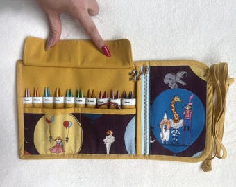 Peut inclure: Un étui à aiguilles à tricoter en tissu jaune moutarde avec un imprimé sur le thème du cirque représentant une girafe, un éléphant et des artistes. L'intérieur contient plusieurs aiguilles en métal de différentes tailles, chacune marquée.