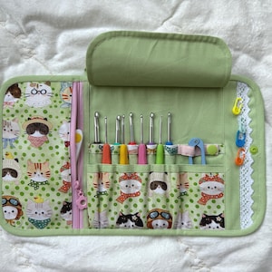 Peut inclure: Un étui de rangement en tissu vert clair pour crochets, orné d'un motif répétitif de chats de dessin animé. L'intérieur comprend plusieurs fentes contenant divers crochets colorés, un outil en plastique bleu et trois marqueurs de mailles.