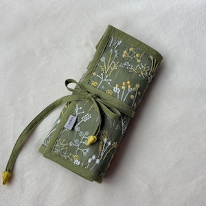 Puede incluir: Un organizador de tela enrollable en color verde oliva con un patr&oacute;n bordado de flores silvestres blancas y amarillas. El organizador se asegura con lazos de tela verde a juego e incluye una peque&ntilde;a etiqueta rectangular con el texto "CASSAN".
