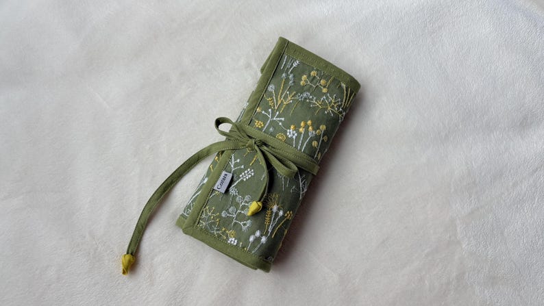 Puede incluir: Un organizador de tela enrollable en color verde oliva con un patr&oacute;n bordado de flores silvestres blancas y amarillas. El organizador se asegura con lazos de tela verde a juego e incluye una peque&ntilde;a etiqueta rectangular con el texto "CASSAN".
