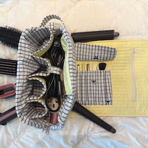 May include: A fabric organizer bag featuring a black and white grid pattern with a pale yellow interior and quilted flap. The bag contains multiple hair styling tools, including brushes and wands, with additional pockets holding small makeup brushes.