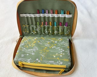 Peut inclure: Un étui zippé jaune moutarde contenant un ensemble d'aiguilles à tricoter en métal coloré organisées dans des fentes étiquetées. L'intérieur présente un tissu vert sauge avec un motif floral blanc et jaune, incluant une pochette zippée.