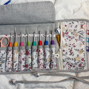 May include: A grey fabric roll-up organiser case featuring a floral interior lining. It contains eight colourful crochet hooks, a pair of light pink scissors, a zipped pocket, and several plastic stitch markers in red, blue, and purple.