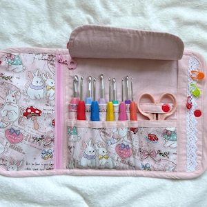May include: A light pink fabric organizer case featuring a pattern of white rabbits, mushrooms, and bows. The interior contains multiple slots holding metal crochet hooks with colorful handles, a pair of light pink scissors, and three plastic stitch markers.