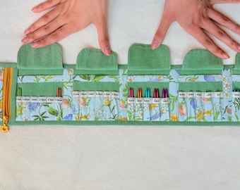 Peut inclure: Un étui de rangement en tissu vert avec un motif floral bleu clair, conçu avec plusieurs poches intérieures et rabats pour contenir diverses aiguilles à tricoter en métal. L'étui comprend une fermeture éclair jaune avec une tirette en forme de fleur.