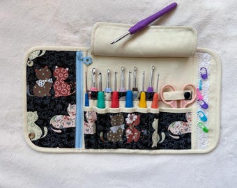 Peut inclure: Un étui en tissu pour crochets avec un motif de chats noirs, contenant plusieurs crochets colorés, une paire de ciseaux rose clair et plusieurs marqueurs de mailles en plastique. Un crochet violet repose sur le rabat supérieur.