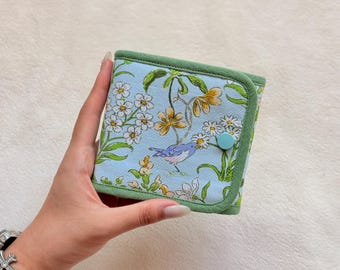 Peut inclure: Un portefeuille carré en tissu avec un fond bleu clair orné d'un motif floral et d'oiseaux dans des tons de jaune, blanc et vert. Le portefeuille est fini avec une bordure en tissu vert uni sur les bords et une fermeture à bouton-pression bleu clair.