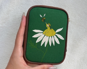 Peut inclure: Une pochette rectangulaire verte à fermeture éclair avec des coins arrondis et une bordure marron. L'avant présente une broderie représentant une personne en robe jaune assise sur une grande marguerite blanche, tendant la main vers une abeille.