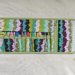 May include: A fabric roll-up organizer featuring a cactus print in shades of green, purple, and white. It includes multiple interior pockets holding colorful crochet hooks and scissors, finished with a white lace trim and green fabric ties.