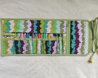 Peut inclure: Un organisateur en tissu enroulable avec un motif de cactus dans des tons de vert, violet et blanc. Il comprend plusieurs poches intérieures contenant des crochets colorés et des ciseaux, avec une bordure en dentelle blanche et des liens.